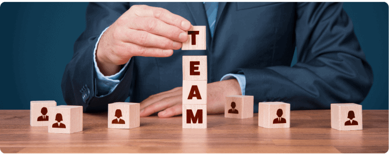 Člověk v obleku skládá kostky s ikonami lidí a nápisem TEAM, symbolizující týmovou práci.
